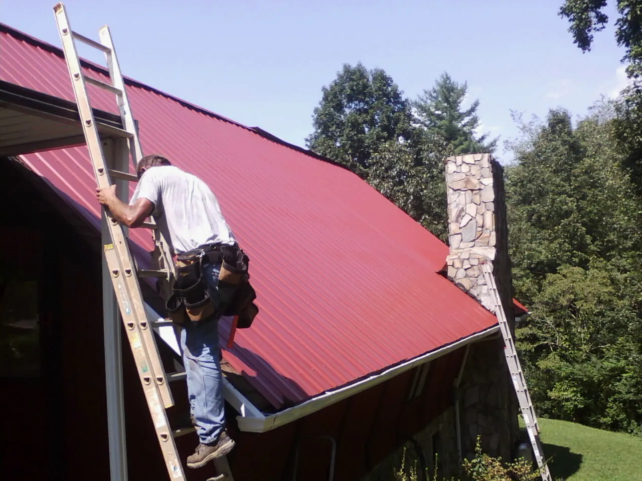 Roofer on a ladder beside a red metal roof and stone chimney — Soffit & Fascia Repair by Roofing Brothers in Uxbridge